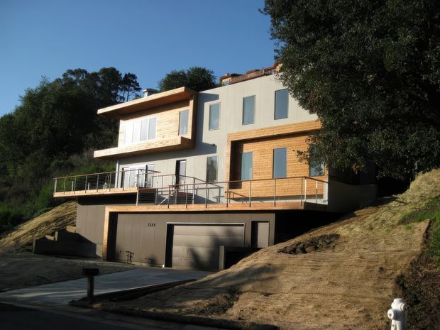 Exterior view of a contemporary multi-level home built into a sloping site, with light rendered walls, warm timber cladding, large glazed openings, a broad balcony, and an integrated double garage. Mature trees frame the property.