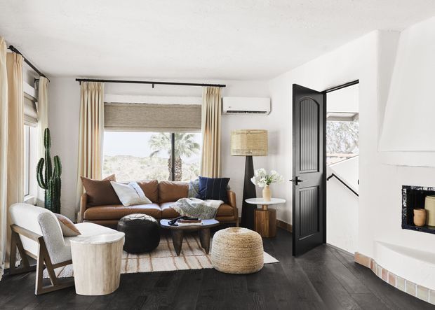 Bright, inviting living room with dark timber flooring, white walls, a brown sofa, woven ottoman, armchairs, a central coffee table, and tall windows dressed with neutral curtains and a woven roller blind. A black paneled door opens to a hallway or stair area, while a curved white fireplace feature occupies the right side. Natural textures, indoor greenery, and neutral furnishings create a relaxed contemporary coastal atmosphere.