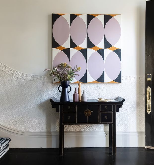 A refined interior vignette featuring a black lacquered console table with drawers and brass hardware, styled with a dark vase, flowers, and decorative ceramic vessels. Above it hangs a large geometric abstract artwork in black, white, pale lavender, and ochre. The room has white textured wall panelling with ornate horizontal moulding, a dark timber floor, and a partially visible traditional black door with brass fittings. A small upholstered seat appears at the lower left.