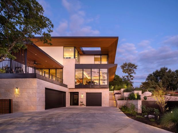 Exterior view of a contemporary multi-level residence at dusk, with broad projecting flat roof planes, warm timber soffits and cladding, extensive dark-framed glazing, pale stone masonry walls, and two integrated garage doors. A paved driveway leads to the house, with landscaped garden beds and retaining walls around the entry approach.