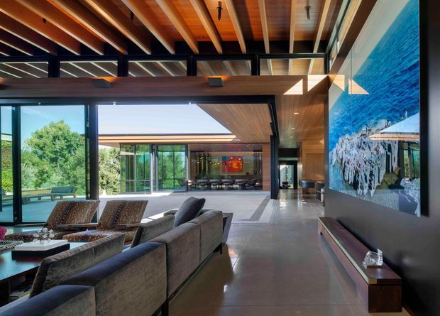 Spacious contemporary living room with expansive floor-to-ceiling glazing opening to a green landscape. The room features exposed warm timber ceiling beams and panels, dark steel-framed windows and doors, polished grey concrete flooring, large upholstered sofas and lounge chairs, and a dramatic oversized blue-toned artwork above a low timber console. A connected glazed living or dining area is visible beyond.