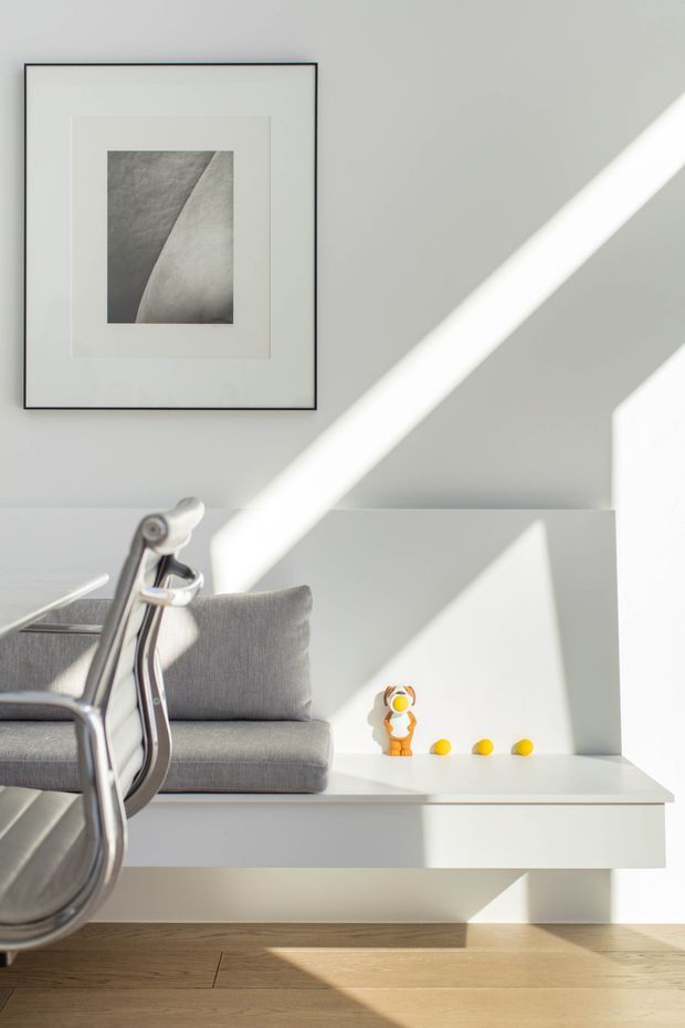 A bright, minimalist interior corner featuring a built-in white window seat or bench with a grey upholstered cushion, beside a chrome-framed chair. A framed monochrome artwork hangs on the light grey wall, while strong diagonal sunlight creates dramatic geometric shadows. The space has a warm timber floor and sparse decorative objects on the ledge.