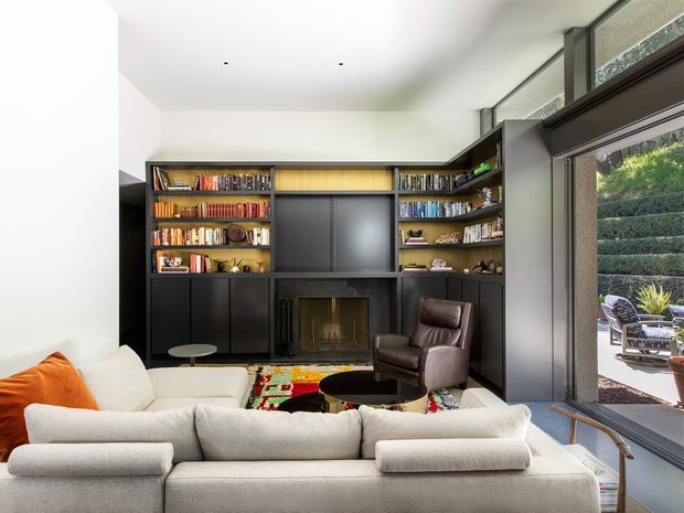 A bright, contemporary living room centered on a full-height dark built-in media and bookcase wall with an integrated fireplace. A large pale sectional sofa faces the built-in, accompanied by a brown leather lounge chair and round dark coffee table. Floor-to-ceiling glazing opens to a landscaped outdoor area, while a colourful patterned rug adds visual contrast.