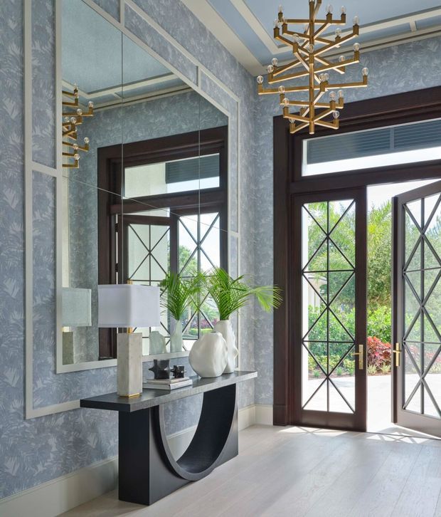 Elegant entry hall with blue-grey patterned wallpaper, dark timber-framed glazed double doors, a large decorative mirror, sculptural black console table, contemporary lamps and ornaments, and a prominent brass-and-glass chandelier. The space is bright and refined, with strong geometric detailing in the door glazing and substantial natural light.