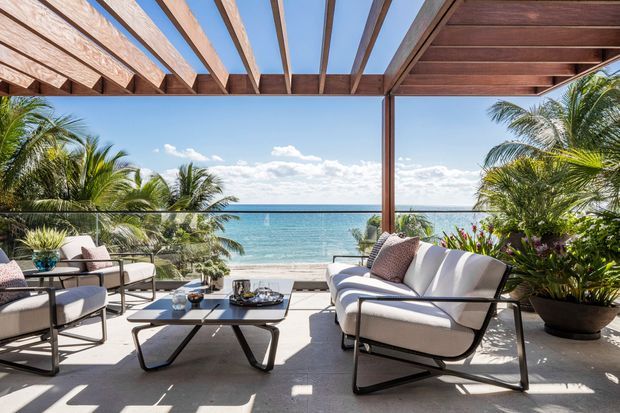 A bright, open-air beachfront terrace overlooking the ocean, furnished with contemporary outdoor lounge chairs, a sofa, and a low coffee table. A timber pergola with exposed slatted rafters provides partial shade, while glass balustrades preserve the panoramic coastal view. Tropical palms and lush potted planting frame the space.