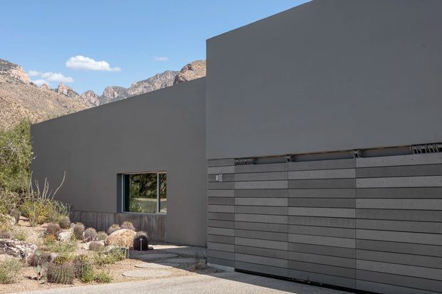 Exterior view of a minimalist, flat-roofed modern building set against rugged mountains. The dark grey rendered facade features a large projecting upper volume, a horizontal slatted garage door, and a small window. A gravel and xeriscaped foreground with desert plants leads to the entry and garage.