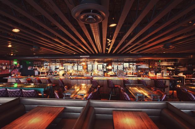 A dark, atmospheric commercial bar and dining venue viewed from the seating area toward a long illuminated back bar. The space features extensive liquor shelving, a continuous bar counter, booth seating, timber dining tables, leather-upholstered banquettes and chairs, and a dramatic exposed timber-slat ceiling with visible ductwork and integrated lighting. The overall fit-out is richly layered, industrial-inspired and suited to a restaurant or cocktail bar.