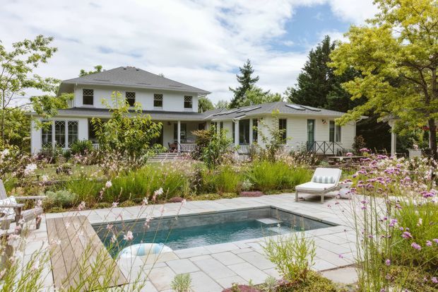 A spacious light-coloured residential house with a low-pitched grey hipped roof, expansive windows, and a covered veranda or terrace. The home is set within a lush, naturalistic garden featuring flowering plantings and a paved patio surrounding a compact rectangular swimming pool. Outdoor lounge furniture is arranged beside the pool, creating a relaxed garden entertaining area.