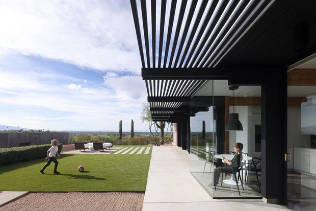 Contemporary single-storey residence with a deep black slatted pergola extending over a paved terrace and full-height glass walls. The outdoor area includes a manicured lawn, low hedges, linear stepping stones, outdoor seating, and sculptural planting, while a dining area and kitchen are visible through the glazing. The image has a strong indoor-outdoor connection, clean horizontal lines, and expansive views toward a distant landscape.