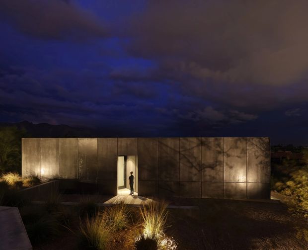 A low, minimalist single-storey building viewed at night, with a monolithic dark metal-clad facade divided into large panels. A narrow illuminated doorway forms the main entrance, while subtle landscape lighting highlights ornamental grasses and the surrounding planted garden. Dramatic storm clouds and a dark silhouetted ridge create a moody setting.