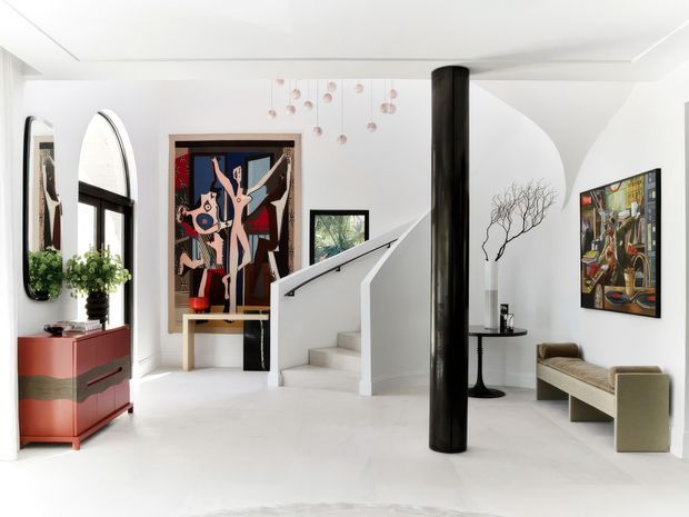 Bright, sculptural residential entry hall with a white open-plan interior, curved staircase and black cylindrical structural column. The space features black-framed arched glazing, a red built-in console, large colourful artworks, a pale upholstered bench, a small round pedestal table, and suspended decorative elements.