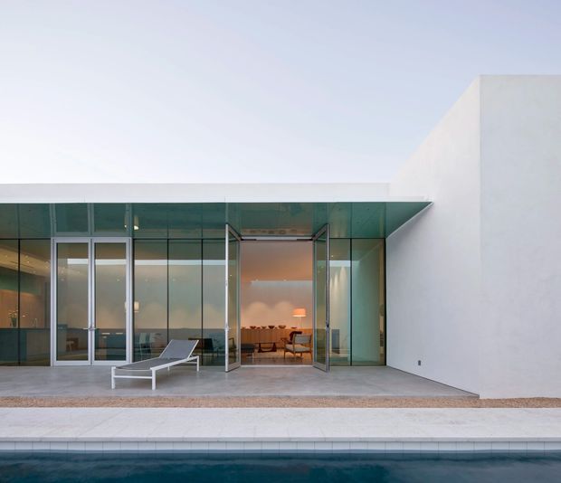 Modern minimalist residence viewed from the pool area, featuring a crisp white flat-roofed volume, extensive floor-to-ceiling glass walls and hinged glazed doors opening onto a pale stone terrace. A dining area is visible inside, while a sleek white outdoor lounge chair sits beside the pool. The composition is clean, geometric and strongly connected to the outdoor space.