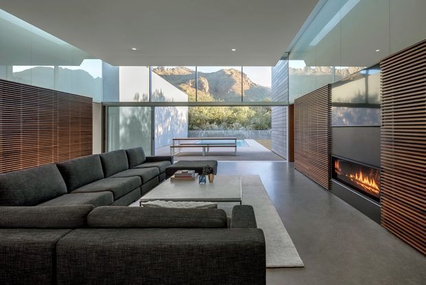 A refined contemporary living room with a large dark sectional sofa, low coffee table, pale area rug, and a linear built-in fireplace. Full-height glazing opens to a mountain landscape and landscaped terrace, while horizontal timber slatted screens line both side walls and conceal integrated storage or architectural elements. The room has a flat ceiling, polished grey concrete floor, restrained neutral palette, and strong indoor-outdoor connection.
