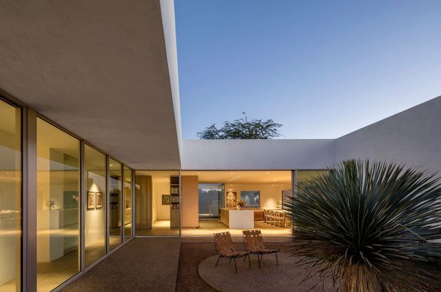 Contemporary single-level courtyard residence photographed at dusk, with broad floor-to-ceiling glazing, smooth concrete or plaster roof and wall planes, warm interior lighting, and a compact landscaped courtyard with sculptural drought-tolerant planting and outdoor lounge chairs. The view looks toward an open living or dining area with a kitchen island beyond.