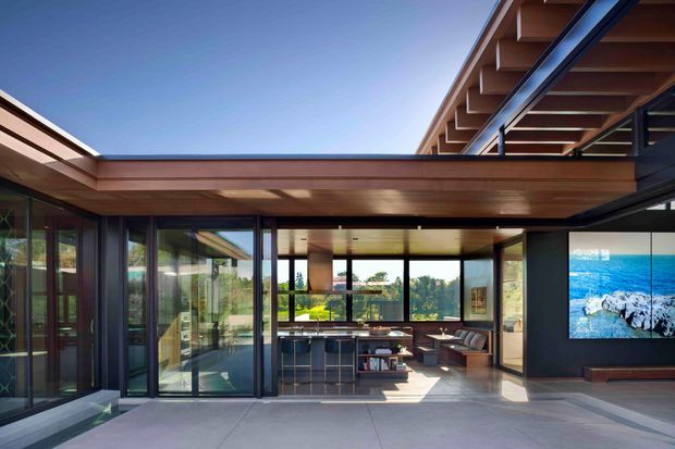 A modern indoor-outdoor living space with extensive floor-to-ceiling glazing, dark-framed glass walls, a timber-lined projecting roof and exposed timber eaves. The open-plan interior contains a kitchen island, dining area and lounge, while a large wall-mounted screen displays a coastal scene. The space opens onto a broad paved terrace and receives abundant natural light.