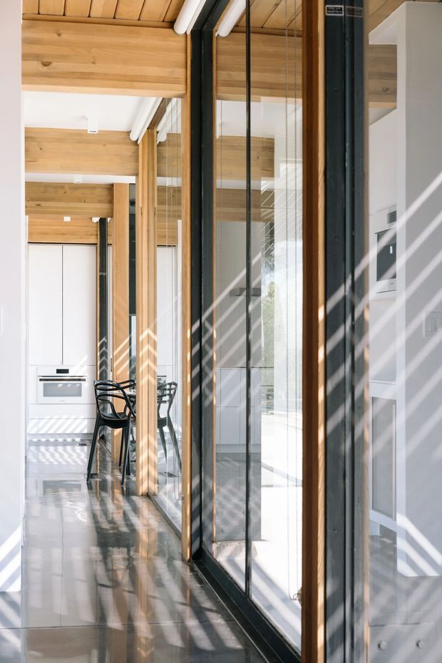 A bright, narrow contemporary interior corridor alongside full-height glazed doors or windows with black metal frames and timber posts. The space opens toward a minimalist white kitchen with an oven and a small dining setting. Exposed timber ceiling beams and timber ceiling lining contrast with white cabinetry, polished light flooring, and strong linear sunlight and shadow patterns across the surfaces.