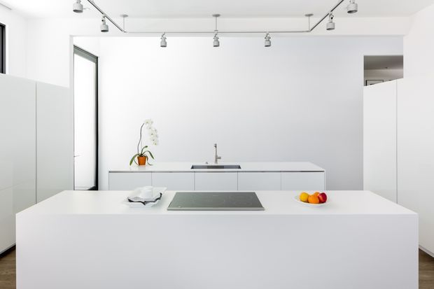 Bright, minimalist white kitchen with a large island, integrated cooktop, sleek white cabinetry, a linear track-lighting system, and a narrow black-framed window. The composition is clean and highly uncluttered, with a small amount of fruit and an orchid providing subtle colour.