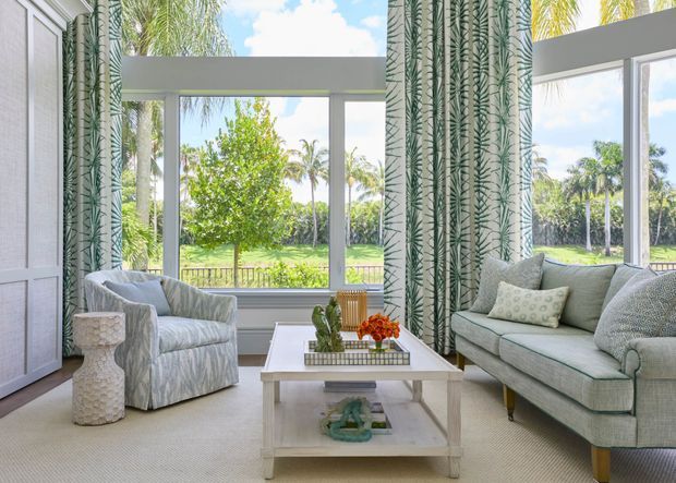 Bright, airy living room with large floor-to-ceiling windows overlooking a lush tropical landscape. The room features pale upholstered seating, patterned botanical curtains, a white coffee table, light carpet, and soft neutral finishes.
