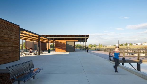 A contemporary low-rise pavilion or community building viewed from a broad concrete terrace. The structure features dark horizontal timber cladding, extensive glazing, a deep projecting flat roof with exposed pergola-like shading, and a dark panelled service door. Timber and metal railings line the terrace, with benches and people using the outdoor space. The setting appears open and suburban, with clear blue sky and distant low-rise buildings.