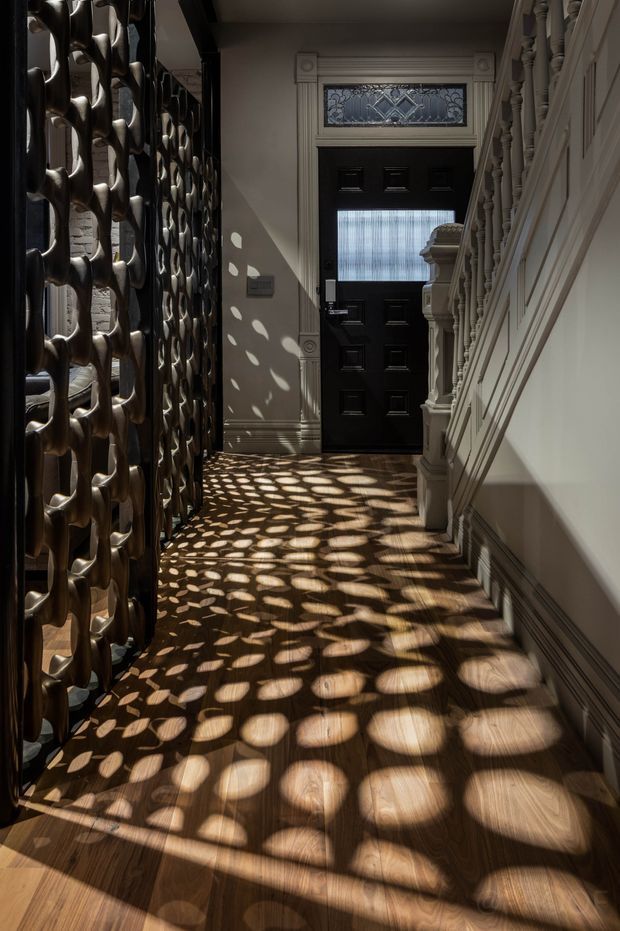 A narrow, elegant residential entry hall viewed toward a black paneled front door with a decorative leadlight transom. The space features polished timber flooring, ornate white wall and stair detailing, a carved balustrade, and a dramatic sculptural dark metal screen on the left casting patterned sunlight across the floor and walls. The architectural detailing gives the hallway a refined heritage character with strong light-and-shadow contrast.