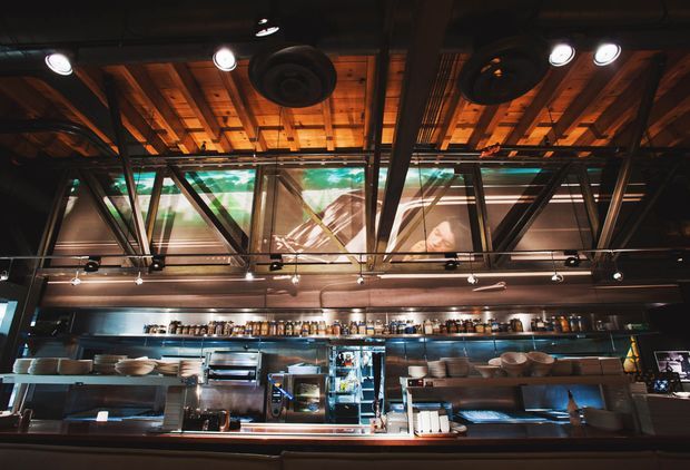 A dark, atmospheric commercial kitchen or bar service area viewed from the customer side. Stainless-steel counters, cooking equipment, shelving, dishes, and beverage containers line the back wall, while a dramatic exposed timber ceiling, black steel beams, track lighting, and large glazed panels create an industrial restaurant interior.