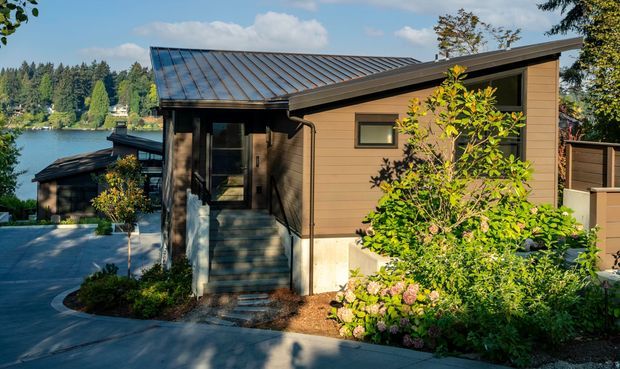 Exterior view of a compact contemporary home on a landscaped, sloping site beside a lake. The building has low-pitched intersecting metal roofs, warm horizontal timber-like cladding, dark-framed glazing, and a covered entry reached by steps. Garden planting and a paved driveway occupy the foreground, with a second lower structure and the lake visible behind.