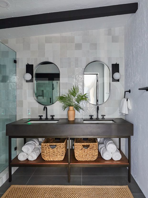 A stylish double-vanity bathroom with a dark metal-framed open console, black countertop, two integrated rectangular sinks, and black tapware. The wall behind the vanity is clad in pale rectangular mosaic tiles, complemented by two arched mirrors and globe wall lights. Woven baskets, rolled white towels, a small plant, dark floor tiles, and a textured woven mat add warmth and contrast. A frameless glass shower enclosure is visible to the left.