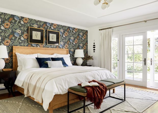 Bright, refined bedroom with a large upholstered bed, layered white bedding, dark blue and green accent pillows, and a botanical floral feature wall. French doors with full-height curtains open to a leafy exterior, while framed artwork, bedside lamps, a bench, and a pale patterned rug complete the room.