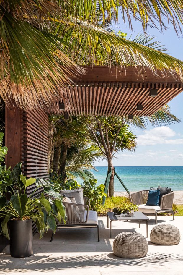 A tropical beachfront outdoor lounge beneath a broad timber pergola with closely spaced slatted rafters. The shaded patio contains woven lounge chairs, a low coffee table, upholstered cushions, textured round ottomans, and lush potted tropical plants. Palm fronds frame views toward turquoise ocean water and a sandy beach under a bright blue sky.
