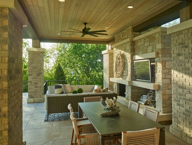 Covered outdoor entertaining area with a dining table and chairs in the foreground, a lounge setting beyond, and a stone fireplace wall with a mounted television. The space has substantial stone masonry, tiled flooring, a timber-lined ceiling with recessed lights, and a large ceiling fan, opening onto a leafy outdoor view.