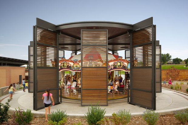 Exterior view of a contemporary open-air pavilion enclosing a carousel, with large dark metal-framed folding screens filled with horizontal timber slats. The circular structure has a curved roof and central slatted screen, set within a landscaped public recreation area with paved paths and low planting.