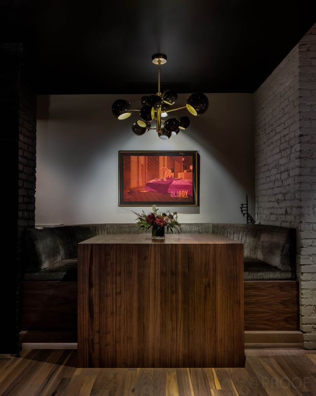 A dark, dramatic lounge or dining area with a built-in upholstered banquette wrapping around the back and sides, centered on a large rectangular dark timber table. The room has black-painted ceiling and walls, exposed white-painted brick side walls, timber flooring, a sculptural brass-and-black globe chandelier, and framed artwork on the rear wall. The overall composition is intimate, moody, and richly textured.