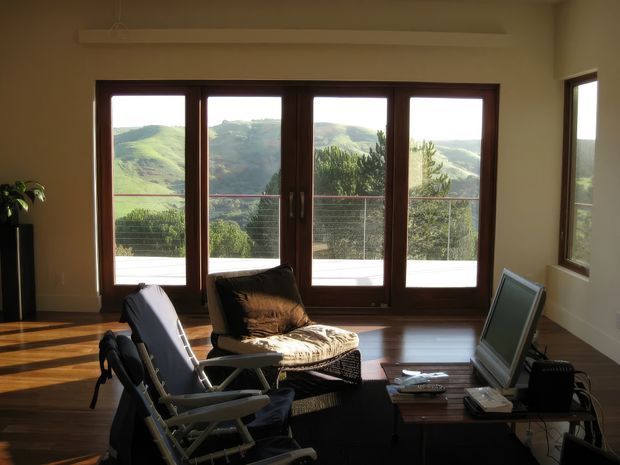 A warm, sunlit living room with dark timber-framed floor-to-ceiling glazed doors opening to a balcony or terrace with expansive green hills beyond. The room contains a lounge chair, woven ottoman, low timber coffee table, and television, with polished timber flooring and neutral walls.