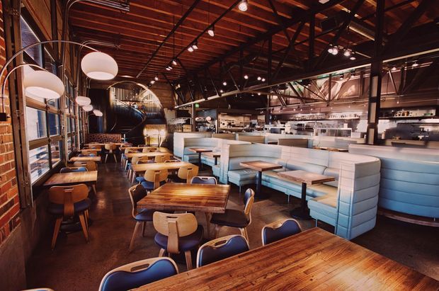A spacious industrial-style restaurant or café interior with rows of timber dining tables and mixed seating, including upholstered banquettes and blue chairs. The space features exposed dark steel framing, a warm timber slatted ceiling, exposed brick walls, pendant globe lighting, polished dark flooring, and an open service counter and kitchen area at the rear.