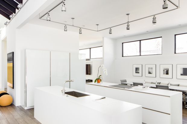 Bright, minimalist open-plan kitchen with a large white island, integrated sink and streamlined white cabinetry. The kitchen opens toward a dining area with a long table and chairs, while high horizontal windows, framed artwork, and a ceiling-mounted track lighting system define the space. Light timber flooring and sparse furnishings create a clean, contemporary interior.