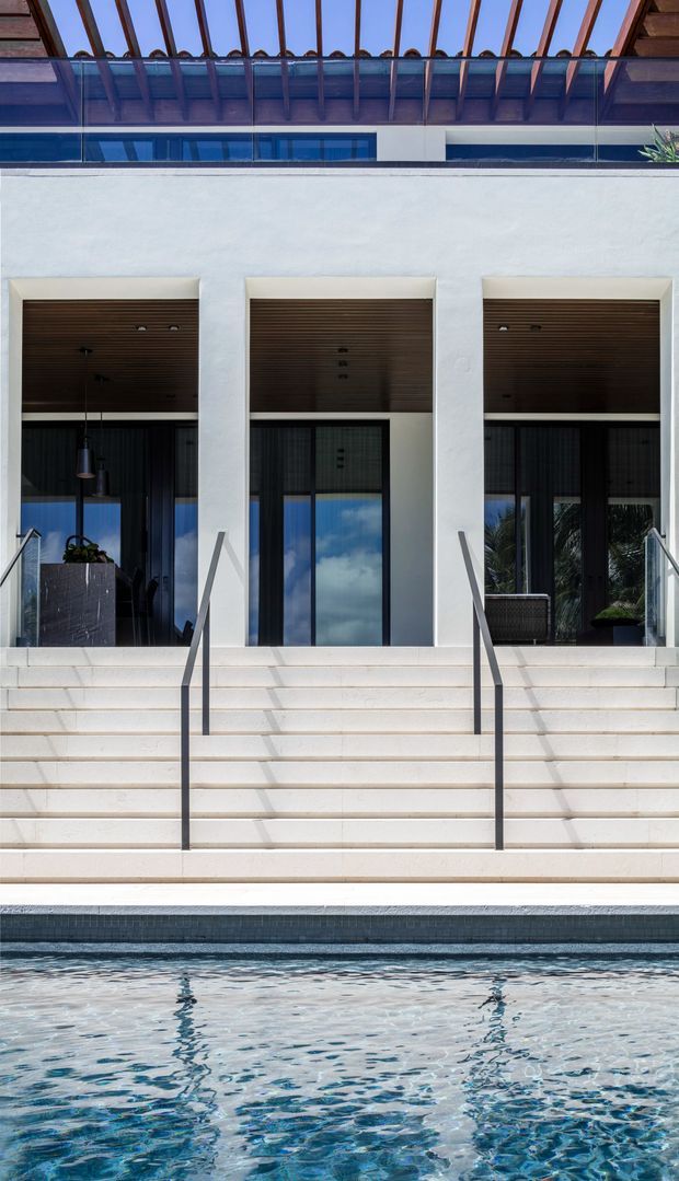 A symmetrical exterior view of a contemporary multi-level residence facing a swimming pool. Broad pale rendered walls frame three recessed glazed openings with dark timber-lined ceilings and large black-framed glass doors. Wide pale steps with slim dark metal handrails lead down to the pool, while an upper terrace features a glass balustrade and exposed timber pergola beams.