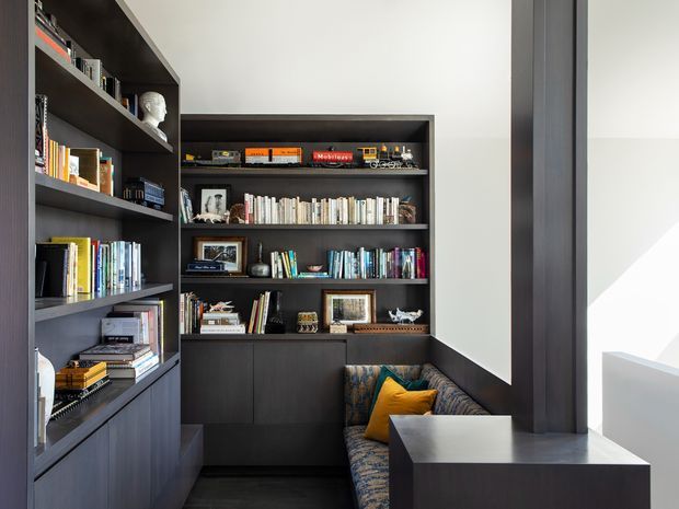A compact, built-in library and reading nook with dark timber shelving and cabinetry wrapping around two walls. The shelves display books, framed photographs, decorative objects and model trains. A patterned upholstered bench seat with mustard and teal cushions sits beside a dark built-in desk or side cabinet, illuminated by strong natural light from the right.