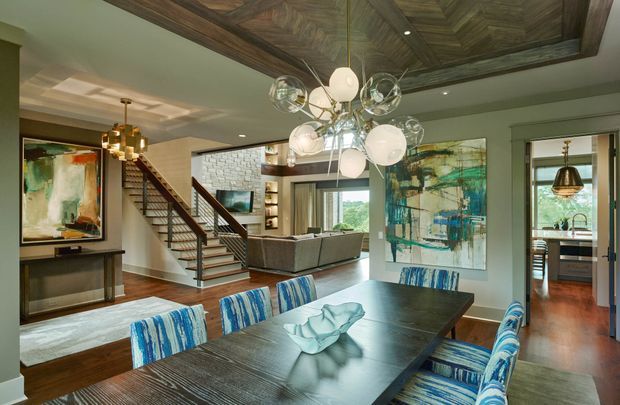 Open-plan dining area with a large dark timber dining table and blue-and-white upholstered chairs in the foreground. The space connects visually to a living room with a sectional sofa, a stairway, and a kitchen beyond. Distinctive fixed architectural details include a richly patterned timber coffered ceiling panel, timber flooring, dark timber stair elements, recessed shelving, and broad interior glazing. Contemporary furnishings are paired with colourful abstract artwork and a sculptural multi-globe pendant light.