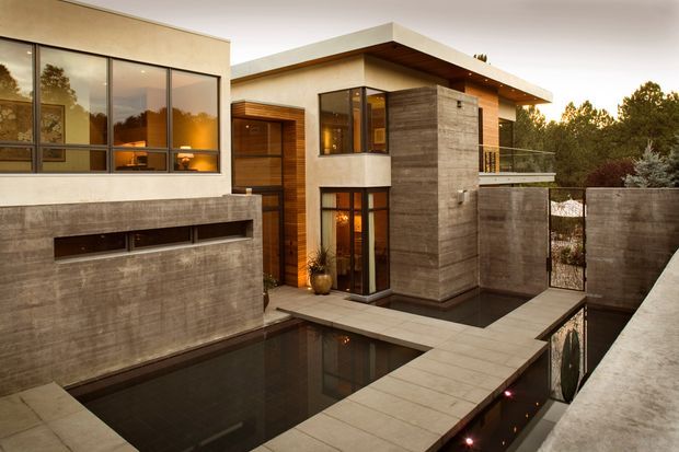 Exterior architectural view of a contemporary multi-level residence arranged around a dark reflecting pool and paved courtyard. The home features broad floor-to-ceiling glazing, flat roof planes, warm timber cladding and soffits, smooth light rendered walls, substantial grey concrete feature walls, and glass balcony balustrades. The scene is photographed at dusk with warm interior lighting and dense trees in the background.