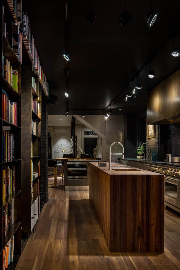 A moody, narrow kitchen interior with a large timber island, dark cabinetry and ceiling, integrated stainless-steel appliances, and a timber floor. Full-height open shelving packed with books lines the left wall, while dark painted brick, timber wall panels, and track lighting create an intimate industrial-library atmosphere. A dining area and entry door are visible beyond the kitchen.