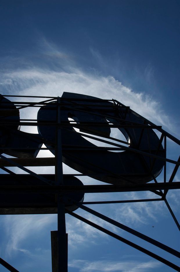 Upward-looking exterior view of a large abstract steel framework supporting dark, curved industrial structures or vessels, silhouetted against a deep blue sky with wispy clouds. The image emphasizes exposed structural members, geometric diagonals, and sculptural industrial form.
