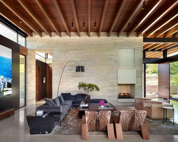 A sophisticated open-plan living room with a dining area, framed by expansive glazing and a dramatic exposed timber-beam ceiling. The room features a pale stone feature wall with a minimalist fireplace, polished concrete flooring, a dark sectional sofa, sculptural timber-framed lounge chairs, and contemporary lighting. Warm timber, natural stone, neutral furnishings, and abundant daylight create a refined yet inviting atmosphere.