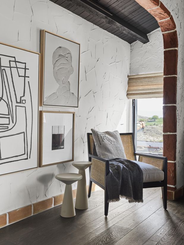 A styled reading corner with a cane-front armchair, grey throw and cushion, and sculptural side tables. The space features heavily textured white plaster walls, dark timber ceiling boards, dark timber flooring, exposed red brick around the window opening, framed monochrome artwork, and a woven roller blind. A rural landscape is visible through the window.