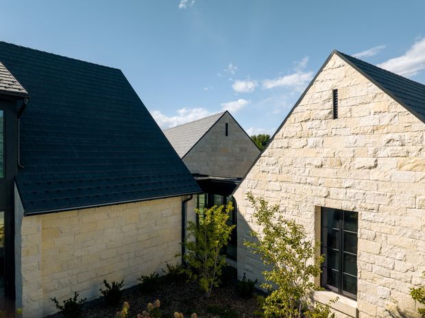 Exterior architectural view of a compact cluster of steeply pitched gabled buildings arranged around a landscaped courtyard. The buildings feature pale rough-cut stone masonry walls, dark metal-looking pitched roofs, and tall narrow black-framed windows. The composition emphasizes the repeated gable forms, courtyard relationship, and strong contrast between light stone and dark roofing under a clear sky.