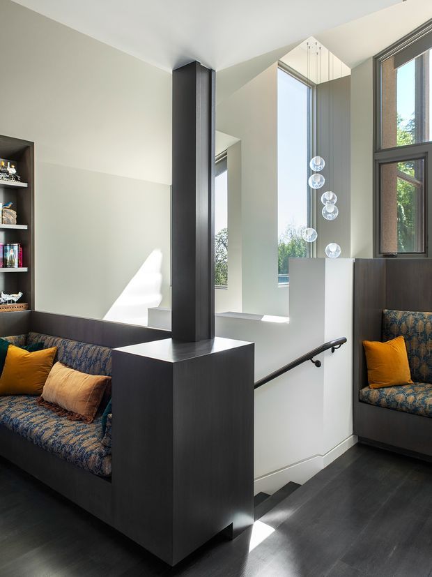 Bright, double-height stair landing and sitting area with dark timber built-in cabinetry and seating, patterned upholstered benches with mustard cushions, tall black-framed windows, a sculptural pendant cluster, and a black handrail descending to the lower level. The space combines strong geometric joinery with abundant natural light.