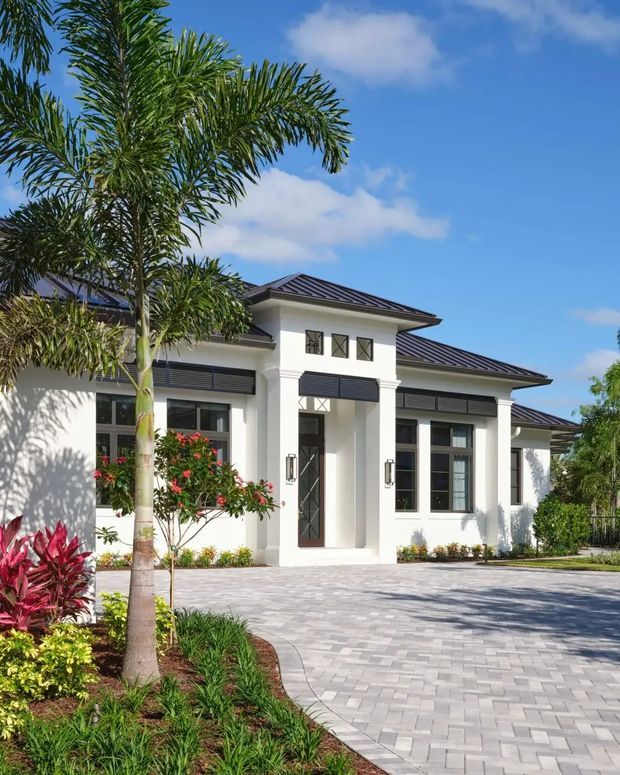 Bright exterior view of a substantial single-storey residence with a symmetrical white rendered facade, dark low-pitched hipped metal roof, columned entry porch, tall gridded windows, and dark louvred or shutter-like sunshade panels. A broad light-grey paved driveway and tropical landscaping with palms and red-leaf plants frame the house.