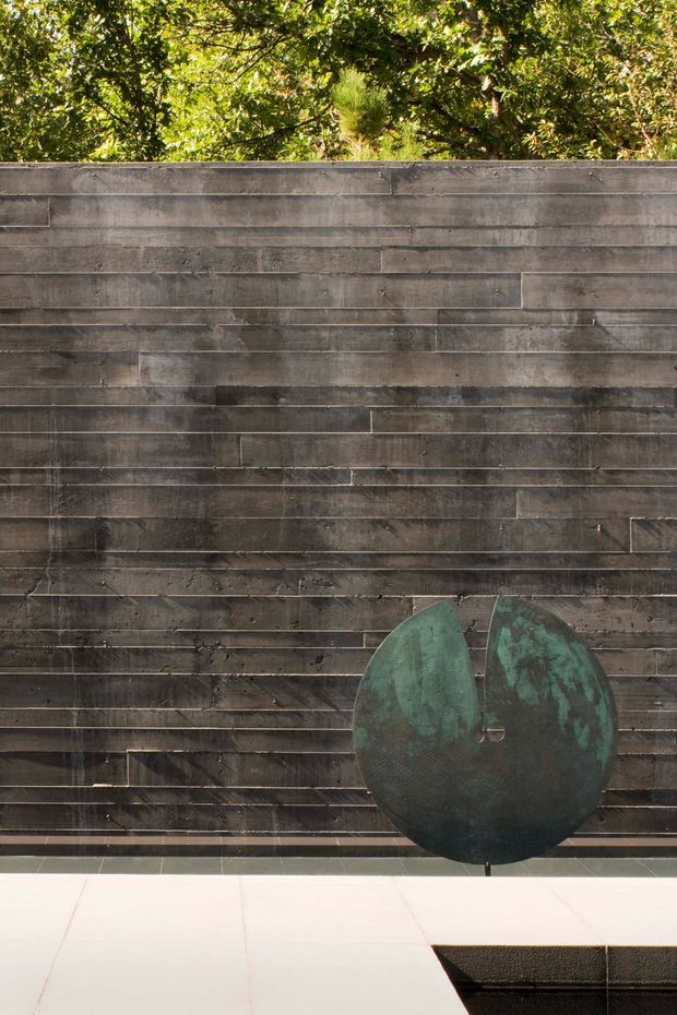 A close-up architectural detail showing a dark, weathered horizontal timber-clad wall with staggered boards. A sculptural oxidised green metal feature is mounted in front of the wall, with leafy planting visible above and pale paving or tiled surfaces at the base.