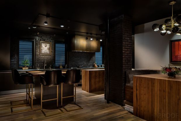 A dark, dramatic open-plan kitchen and dining area with exposed dark brick walls, timber cabinetry and flooring, black countertops, and black bar stools with brass-toned frames. Track lighting and a sculptural globe pendant provide focused illumination. A central brick structural pier separates the kitchen from a dining or living zone with a timber sideboard and framed artwork.