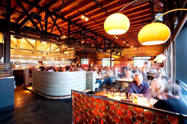 A busy, warmly lit contemporary restaurant interior with exposed timber ceiling boards, black steel trusses and beams, exposed brick walls, large glowing pendant lights, and an open kitchen or service counter. Booth seating and dining tables fill the space, while a prominent front counter features a glossy circular-patterned red-orange decorative surface. The image has a lively hospitality atmosphere with strong industrial architectural detailing and warm amber lighting.