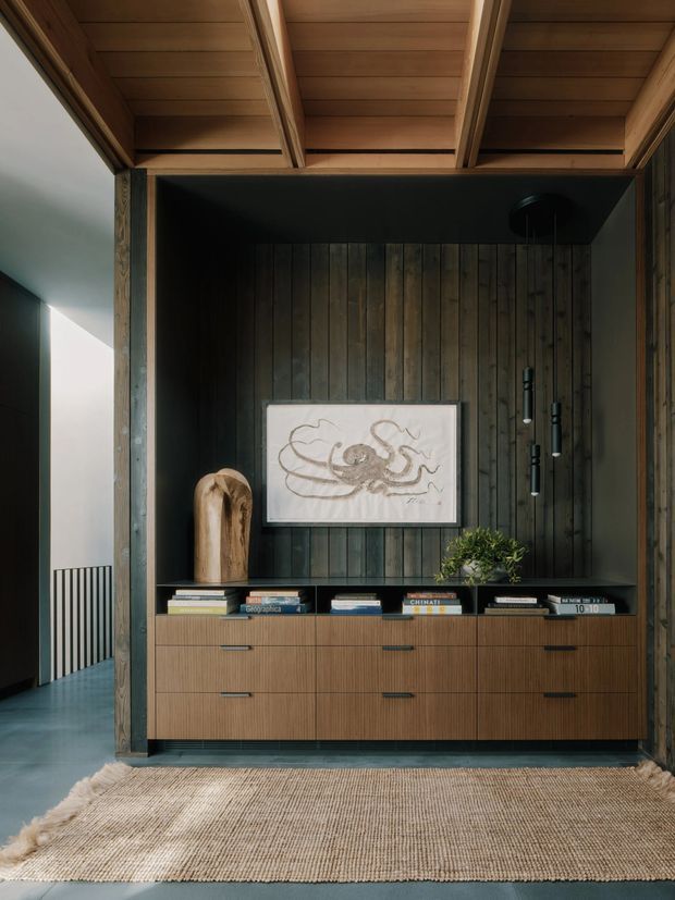 A refined built-in timber media and storage wall in a dark-lined alcove, featuring vertical wood panelling, a low fluted timber cabinet with drawers, display shelves, framed artwork, books, and a small plant. Exposed timber ceiling beams and a woven neutral rug add warmth, while a glimpse of a stair or hallway is visible at left.